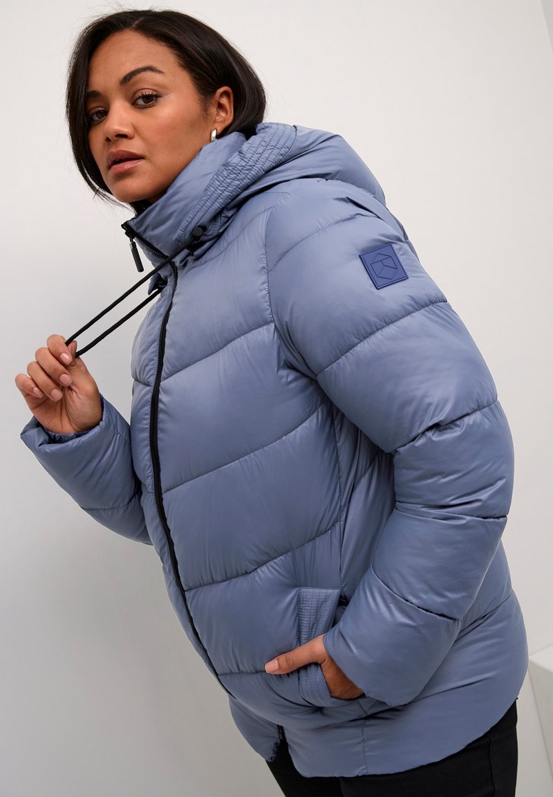 Veste matelassée bleu clair, dotée d'une capuche, de cordons de serrage, d'une fermeture éclair à l'avant et de poches latérales. Texture lisse avec un design matelassé.