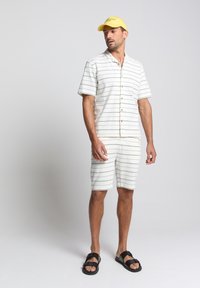 Witte korte mouwen overhemd met lichtgrijze strepen, gecombineerd met bijpassende shorts. Set compleet met zwarte sandalen en een gele pet.