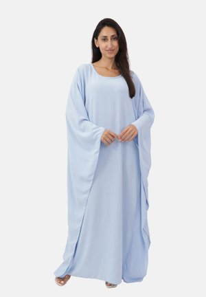 Mujer con el cabello largo y oscuro que lleva un vestido azul claro, suelto y fluido, de pie contra un fondo liso y claro.