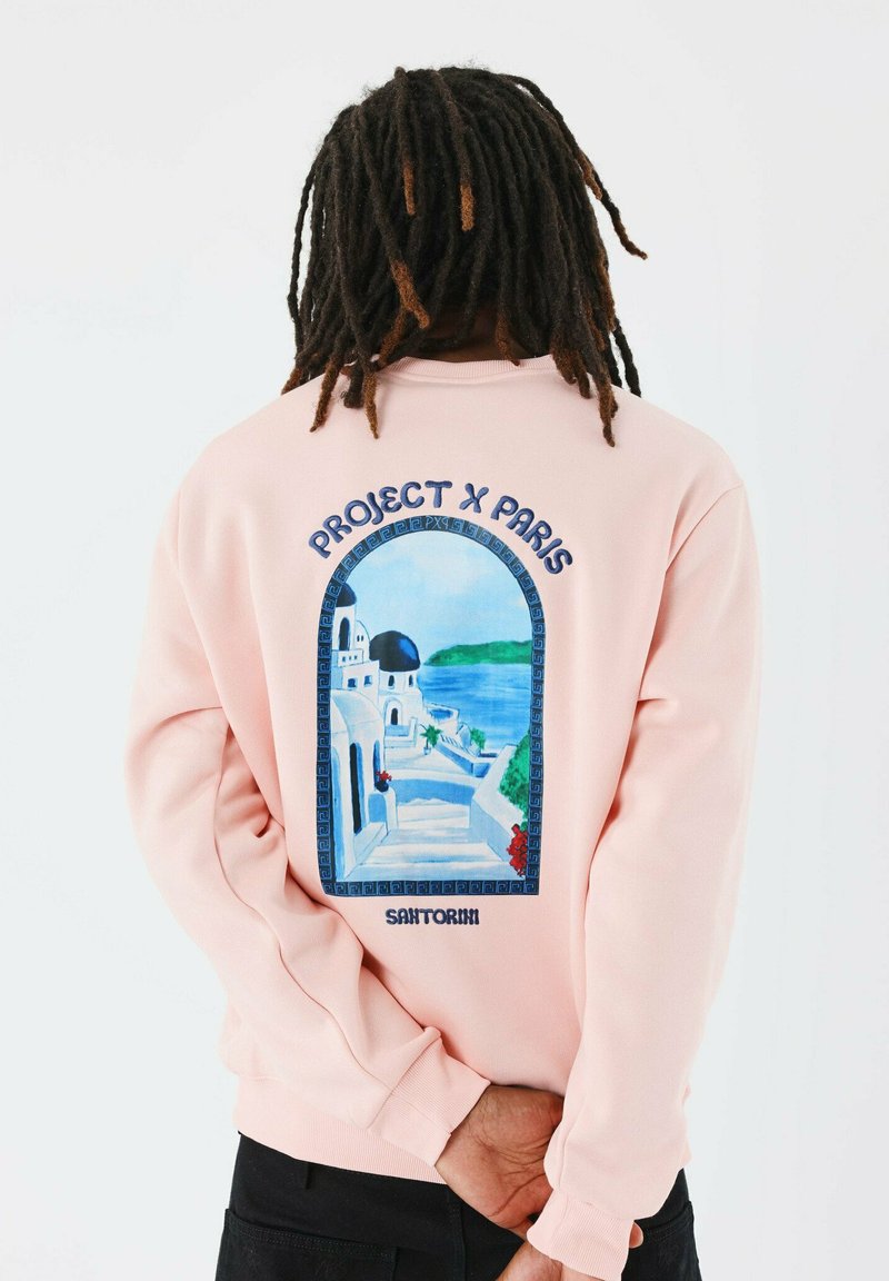 Sudadera de color rosa claro con un gráfico en la parte posterior de una escena de Santorini en azul y blanco, con el texto "PROJECT X PARIS" y "SANTORINI".