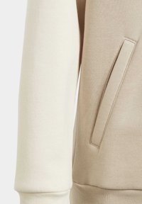 Bomullströja med en krämfärgad vänsterärm och en beige högersida, samt en framficka, slät textur och ribbade muddar.