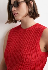 Frau mit kurzen braunen Haaren, die eine Schildpatt-Sonnenbrille, silberne Creolen und ein ärmelloses rotes Strickoberteil trägt.