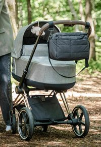 Grå stofklapvogn med en sort base, udstyret med et brunt håndtag, en aftagelig sort opbevaringspose og fire hjul på en skovbund.