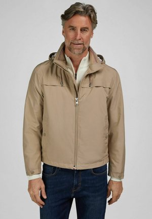 Uomo di mezza età che indossa una giacca beige con zip sopra un maglione bianco e jeans blu scuro, in piedi davanti a uno sfondo uniforme.
