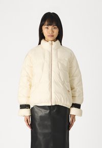 Giacca puffa crema con colletto alto, chiusura con zip e orlo elastico. Presenta polsi neri con dettagli del logo e un design trapuntato.