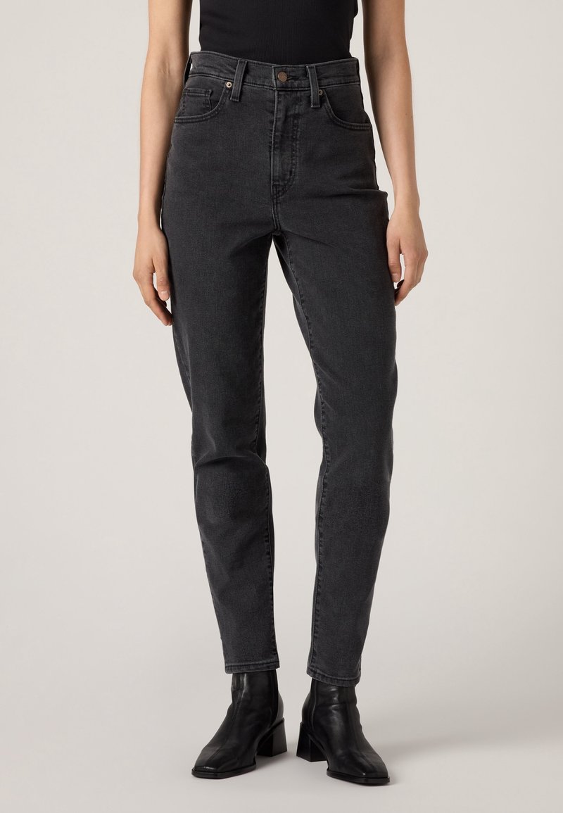 Femme portant un jean noir taille haute, délavé, coupe droite, avec des bottines noires à talons et un haut noir sans manches.