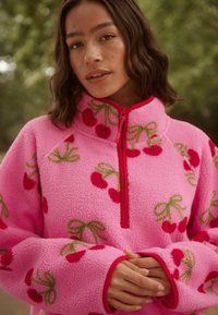 Fleece-Pullover in leuchtendem Pink mit rohem Reißverschluss, mit Kirsch- und Schleifenmuster in Rot und Grün. Weicher Stoff und lockere Passform.