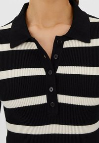 Polo shirt à rayures noires et blanches en ribbed avec col, présentant une patte de boutonnage et un tissu texturé. Les boutons sont circulaires et noirs.
