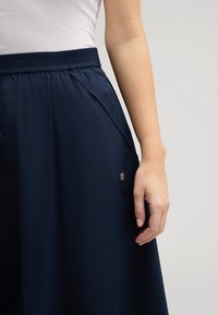 Marineblaue Rock mit elastischem Bund und seitlichen Taschen. Mit einem goldenen Akzentdetail am Saum. Glattes, leichtes Material.