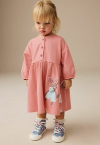 Rosa, strukturierte Kleid mit langen, gepufften Ärmeln und Knopfleiste. Mit einer Stickerei eines Kaninchens, das einen Regenschirm hält. Getragen mit gemusterten Sneakers.