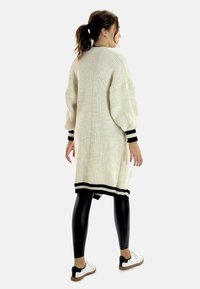 Lång, krämfärgad stickad cardigan med ett texturerat mönster, svarta ränder vid ärmsluten och nederkant, kombinerad med glänsande svarta leggings och vita skor.