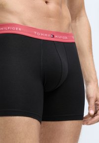 Mustat bokserit, joissa on vaaleanpunainen jousto vyötäröllä ja "TOMMY HILFIGER" -brändäys. Kangas näyttää sileältä ja mukautuvalta.