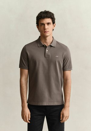 Giovane uomo con capelli corti, ricci e scuri, indossa una polo marrone a maniche corte e pantaloni neri, in piedi davanti a uno sfondo chiaro e uniforme.