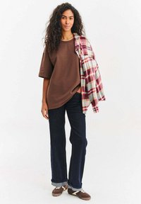 Hnědé tričko volného střihu, doplněné tmavě modrými džíny s širokými nohavicemi. Červeno-bílé kostkované košile přehozené přes jedno rameno; hnědé boty s bílými detaily.