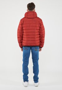 Giacca puffa trapuntata rossa con cappuccio, caratterizzata da cuciture orizzontali e una forma leggermente aderente. Abbinata a jeans blu e sneakers bianche.