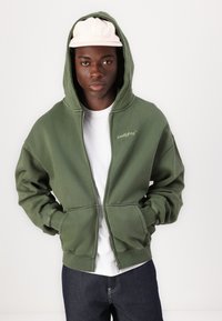 Olivgrüner Reißverschluss-Hoodie mit einer vorderen Tasche, gerippten Bündchen und einem weißen Logo. Über einem weißen T-Shirt getragen. Hellrosa Mütze auf dem Kopf.