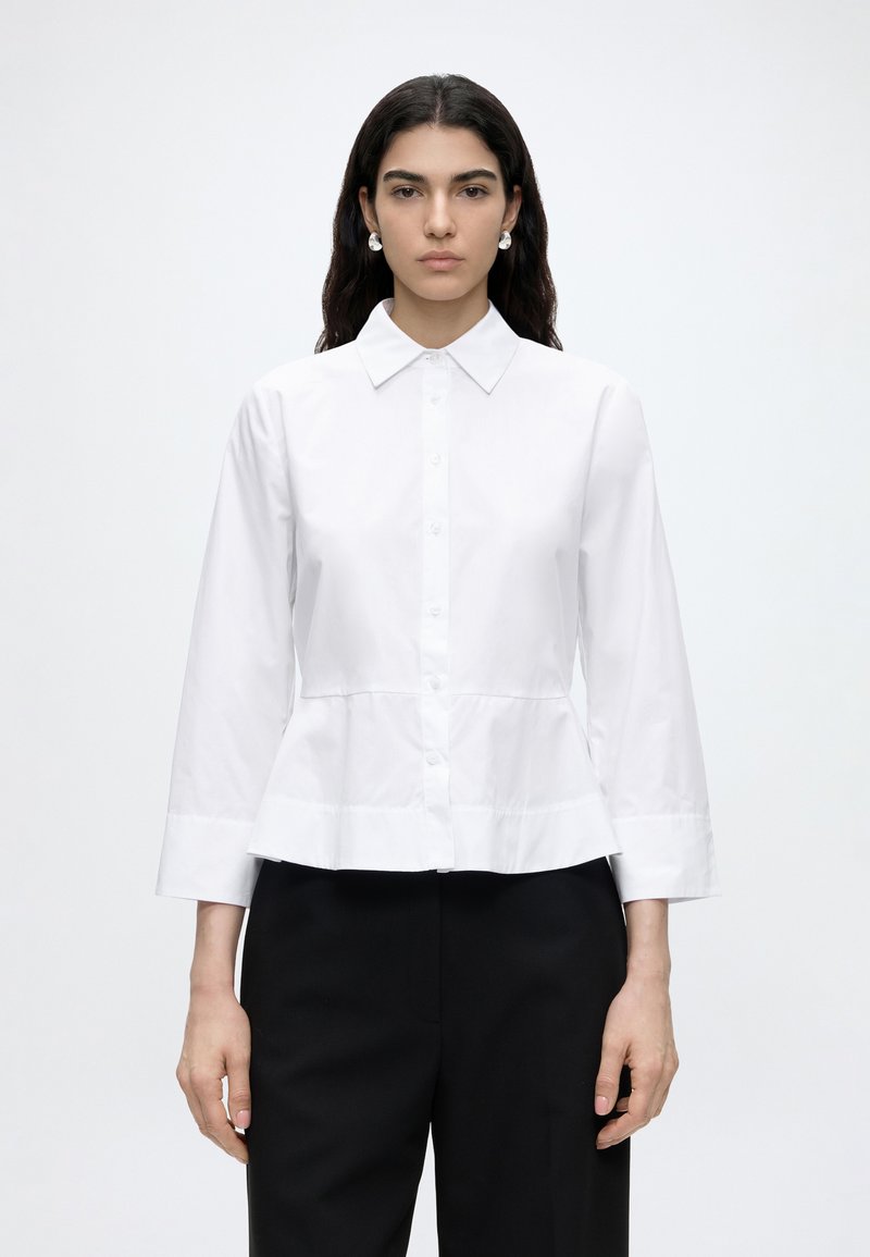 Femme aux longs cheveux foncés portant une chemise blanche boutonnée à manches longues et un pantalon noir, debout devant un fond clair uni.