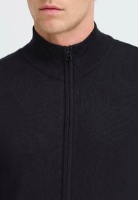 Svart, zip-up tröja med hög ribbad krage, som har en slät textur och minimala sömmar för en ren, modern look.