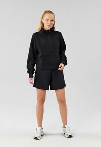 Svart fleece zip-up hettegenser og matchende shorts, med høy krage, framlomme og slitesterk stoff, båret sammen med hvite joggesko.