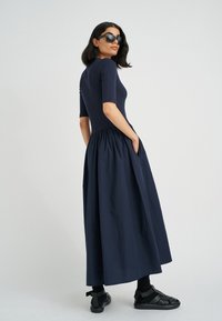 Dunkelblaues Kleid mit einem taillierten, gerippten Oberteil und einem weiten Rock. Mit kurzen Ärmeln und seitlichen Taschen. Kombiniert mit schwarzen Sandalen.