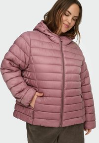 Kvinne iført en lilla, quiltet dunjakke med hette, stående med den ene hånden i lommen, foran en ensfarget bakgrunn.