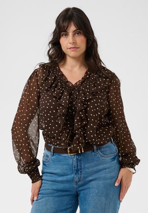 Femme debout, portant un chemisier marron transparent à manches longues avec des pois blancs et des volants, et un jean bleu avec une ceinture marron. Fond neutre.