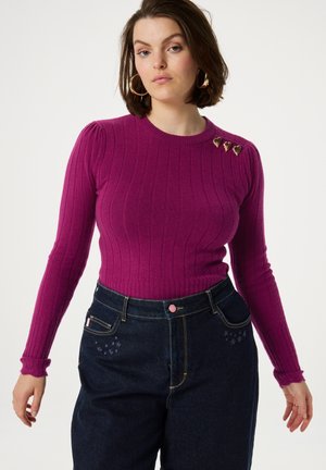 Fuchsia ribbeltrui met lange mouwen, een ronde hals en drie gouden hartvormige versieringen op de linkerschouder. Donkerdenim high-waisted broek.