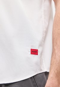 Λευκό πουκάμισο που φοράει ένα άτομο, με μια ορατή κόκκινη ορθογώνια ετικέτα που γράφει "HUGO" κοντά στο κάτω στρίφωμα και εμφανίζεται μέρος του δεξιού μανικιού.