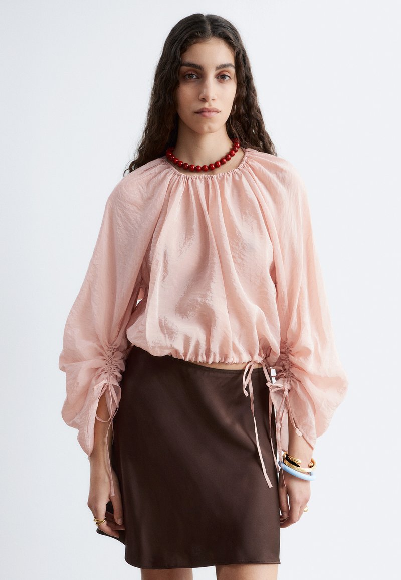 Femme aux longs cheveux foncés portant un chemisier rose clair brillant froncé, une jupe marron foncé, un collier rouge en perles et des bracelets sur fond blanc.