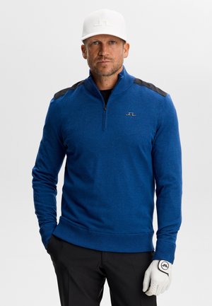 Hombre con un suéter azul con cierre hasta el pecho, gorra blanca, pantalones negros y un guante de golf blanco, de pie con las manos en los bolsillos sobre un fondo liso.