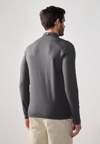 Polo de manga larga en algodón gris oscuro, con un cuello y un pequeño acento rojo y blanco en el escote. Textura suave, diseño ajustado.