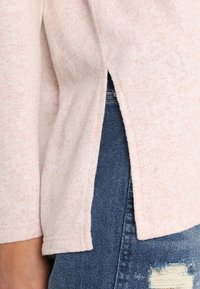 Ljusrosa tröja med mjuk textur, med sidospalter och ärmar i trekvarts längd, lagd över blå slitna jeans.