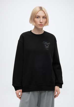 Personne blonde portant un sweat-shirt noir oversize avec un papillon et le texte « Maybe this is paradise » sur la poitrine, associé à un pantalon gris.