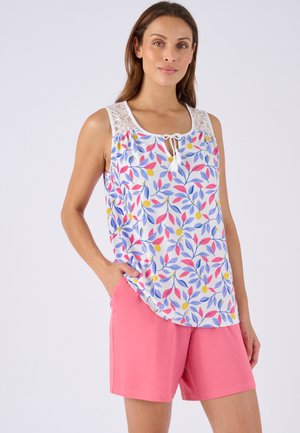 Femme portant un haut fleuri sans manches avec des détails en dentelle et un short rose, debout avec une main dans la poche sur un fond uni.