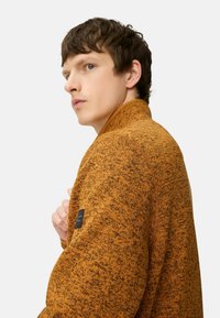 Strikket orange-brun sweater med krave, der har et sort logo-patch på ærmet og et strikket mønster overalt.