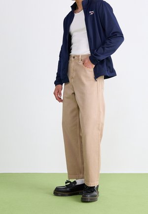 Chaqueta de zip navy, camiseta sin mangas blanca, pantalones anchos beiges, zapatos negros con cordones y calcetines blancos, todo sobre un fondo claro.