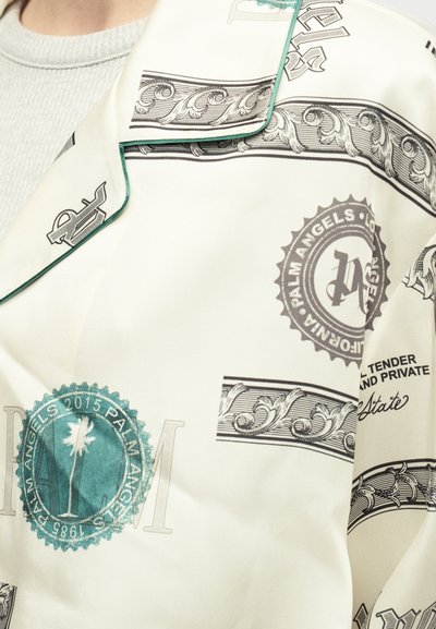 Μεταξωτό πουκάμισο με κρεμ βάση, που διαθέτει μαύρα και πράσινα μοτίβα, λογότυπα και διακοσμητικά στοιχεία. Επιπεδος γιακάς και λεία υφή.