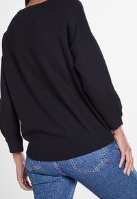 Schwarz langärmliges Oberteil aus strukturiertem Stoff, mit lockerer Passform und weitem Halsausschnitt, kombiniert mit blauen Jeans aus Denim mit Details an den Hintertaschen.