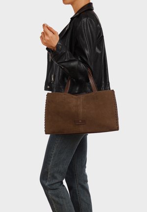 Persona con giacca di pelle nera e jeans scuri che porta una grande borsa tote in suede marrone con bordi cuciti e doppi manici.