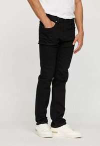 Homme portant un jean slim noir, des baskets blanches et un t-shirt blanc, avec une main dans la poche, sur un fond uni.