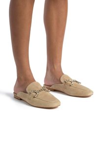 Mocassini beige in suede con punta rotonda e un dettaglio decorativo a catena con strass sulla parte anteriore. Suola piatta, design minimalista.