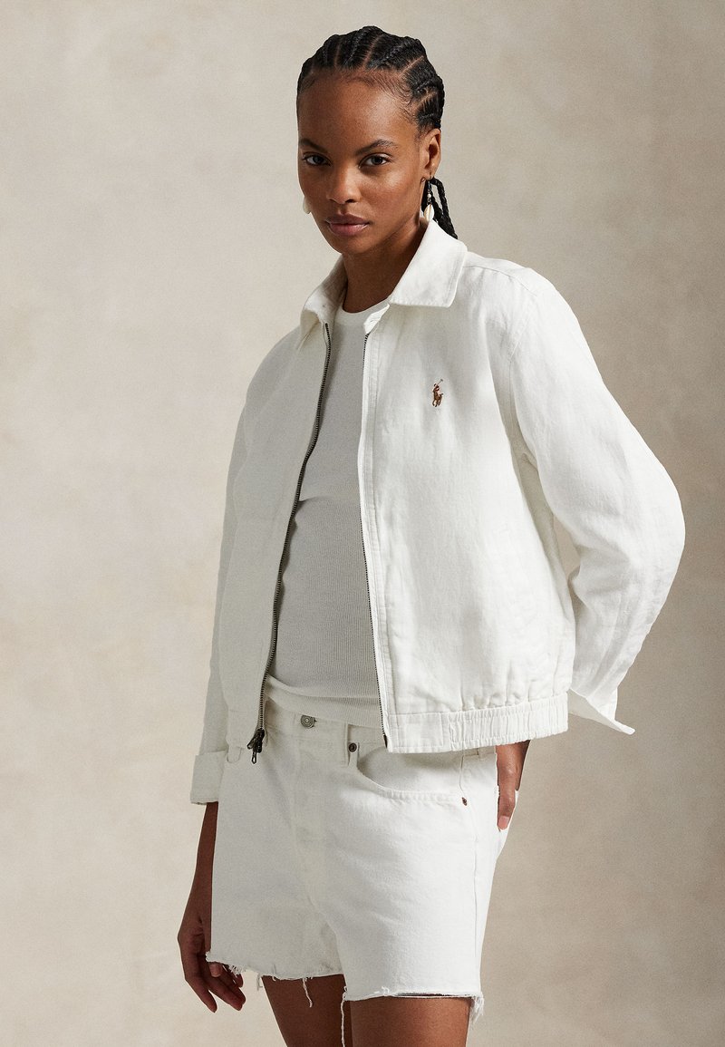 White denim jacket with a zip closure, banded cuffs, and a subtle logo. Worn over a light top and paired with white denim shorts.