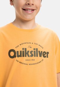 Keltainen puuvillapaita, jossa on musta painettu logo Quiksilverille, kaarevalla tekstillä ja pienemmillä yksityiskohdilla alla, rentoa istuvuutta.