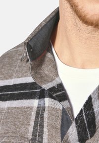 Grau kariertes Hemd mit Kragen, das schwarze und weiße Streifen aufweist. Aus strukturiertem Stoff gefertigt, mit kontrastierendem Innensaum am Kragen. Weißes Hemd darunter.