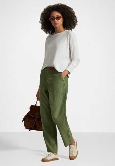 Pantaloni cargo verdi con tasche laterali, abbinati a una camicia bianca a maniche lunghe e scarpe da ginnastica color crema. Gli accessori includono una borsa marrone e occhiali da sole rosa.