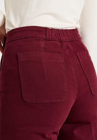 Pantalones de pana color burdeos con una cinturilla elástica, que cuentan con dos bolsillos traseros y un detalle de costura visible en la cintura y los bolsillos.