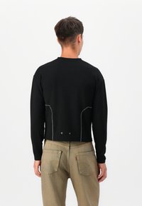 Sort cropped struktureret sweater med lange ærmer, der har kontrasterende hvid syning og to dekorative knapper bagpå.