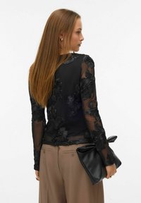 Kvinde med langt brunt hår iført gennemsigtigt sort bluse med blomsterbroderi, beige bukser, og holder stor sort læderclutch.