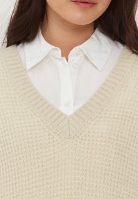 Pull en V couleur crème en tricot texturé superposé sur une chemise blanche à boutons, avec un ourlet côtelé et un col structuré.