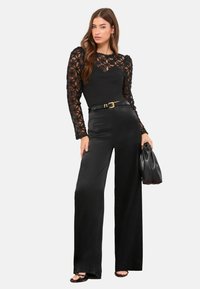 Juodas kombinezonas su ilgomis permatomomis nėrinių rankovėmis, puoštomis gėlių raštais. Šilkinio pojūčio audinys, plačios kelnės, liemuo pabrėžtas juodu diržu.